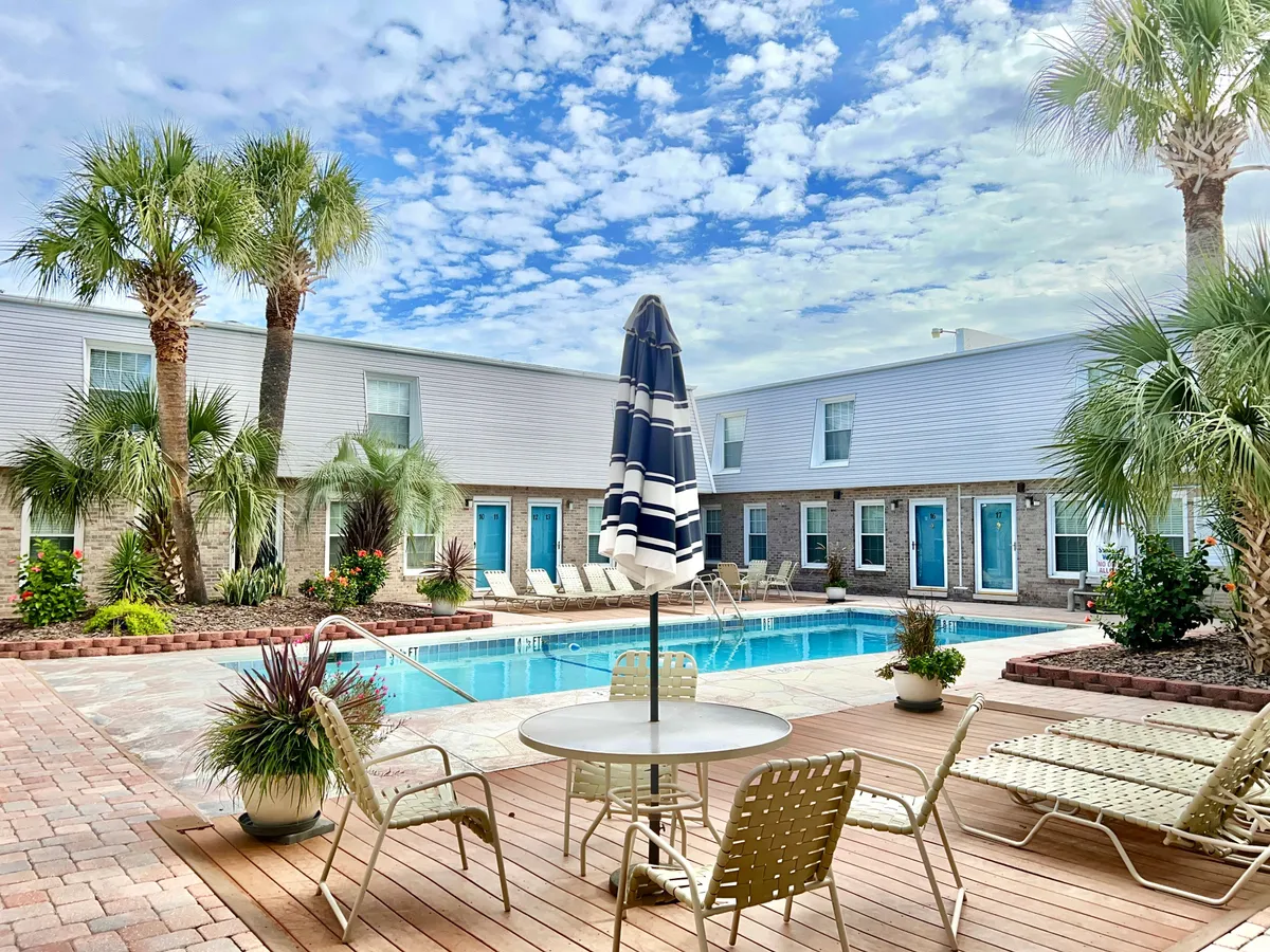 The image features the sparkling swimming pool surrounded by comfortable seating, shaded umbrellas, and lush palm trees. The setting is casual and cheerful, with golden sunlight suggesting either a sunrise or sunset, inviting guests to relax, swim, or socialize in the warm North Myrtle Beach ambiance.