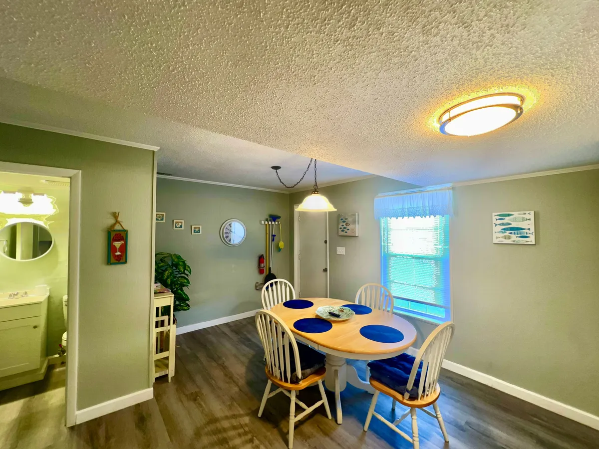 A charming dining area with a table set with blue placemats, a round mirror reflecting light, airy curtains on windows, and decorative wall art. The mood is cheerful yet intimate, ideal for meals, conversations, and casual gatherings.