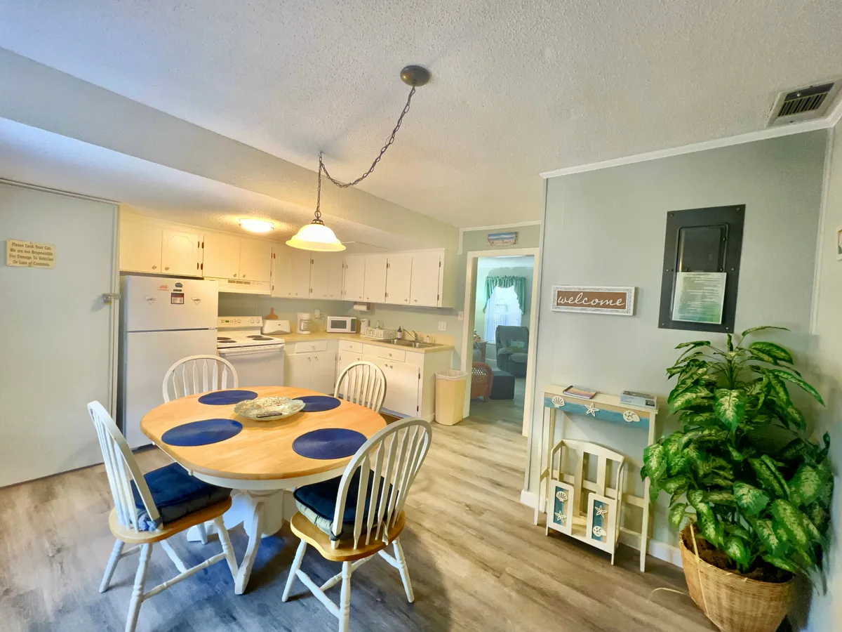 This image features a dining table set within view of the kitchen area, with visible modern kitchen appliances, a welcoming 'welcome' sign, and a potted plant. The scene hints at a cozy space ideal for family meals, celebrations, and intimate gatherings.
