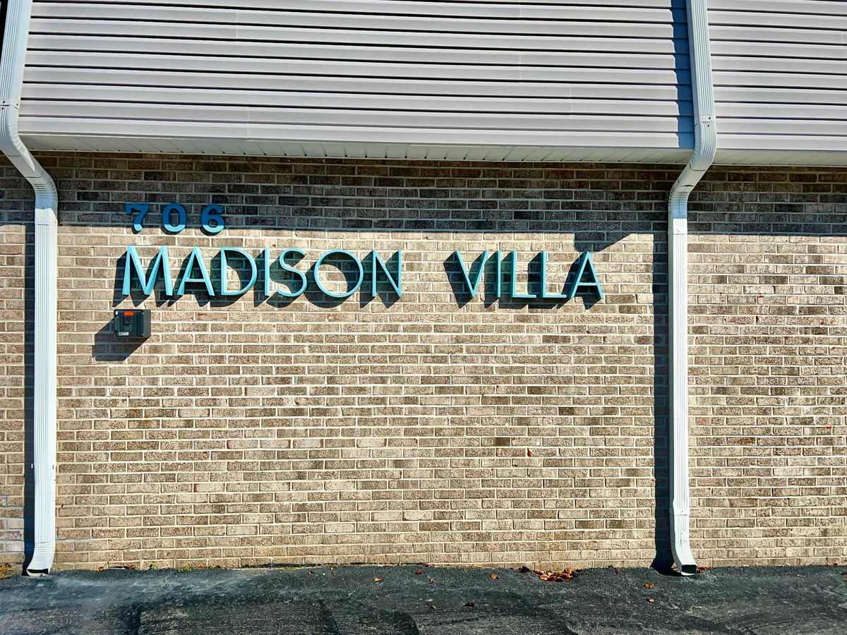 A welcoming exterior image featuring the Madison Villa sign set against a brick wall, complete with a warm light fixture. This entryway scene sets the tone for a friendly and inviting community experience.