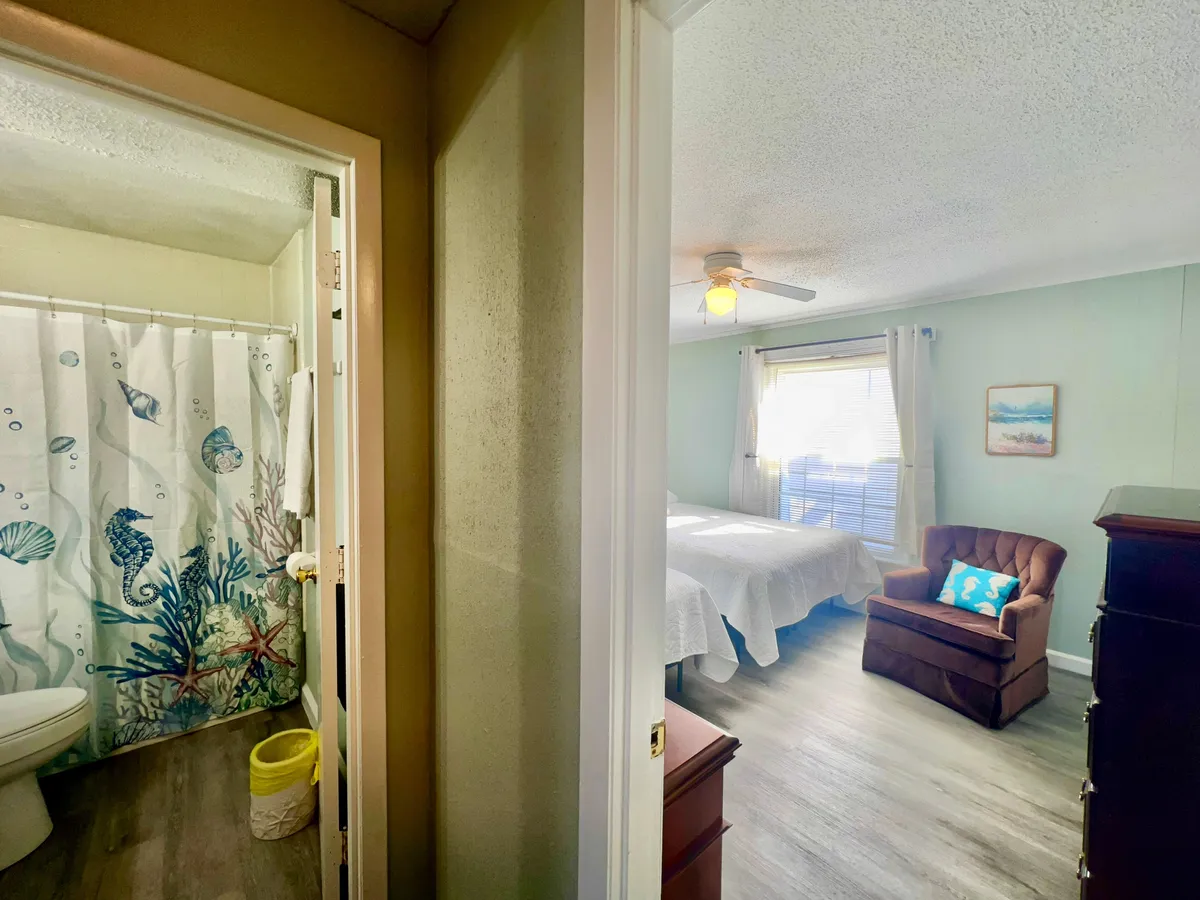 This image features a light-filled bedroom with multiple windows, a cozy armchair adorned with a decorative pillow, and a vibrant shower curtain with a marine life pattern visible in the background. The abundance of natural light streaming indoors evokes the feeling of waking up to the fresh coastal morning, while the subtle beach-themed decor hints at the villa’s proximity to the shoreline.