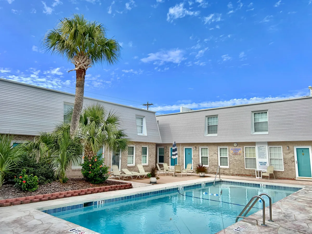 An exterior shot of the community pool area, framed by swaying palm trees and lined with lounge chairs. The building’s facade gives context to the property, while the tranquil pool promises space for both relaxation and playful moments.