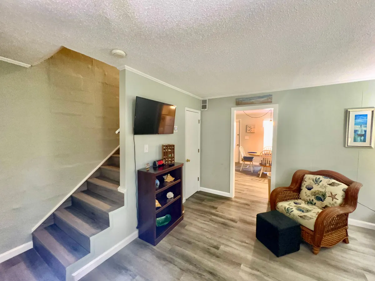 A welcoming living space featuring a cozy rattan chair, a staircase suggesting connection to other rooms, a TV for entertainment, and a decorative shelf. The setting is casual and inviting, ideal for socializing or relaxing after a day out.